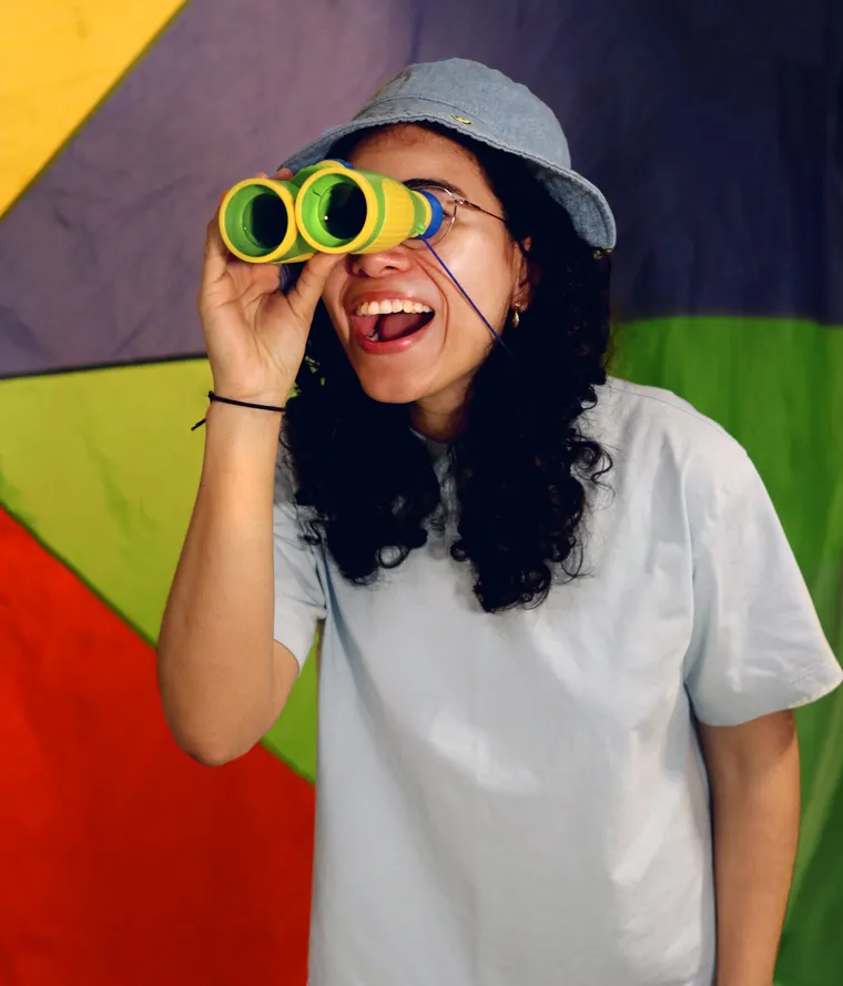 Karla, wearing a denim bucket hat, laughs as she looks through toy binoculars in front of a colorful parachute.
