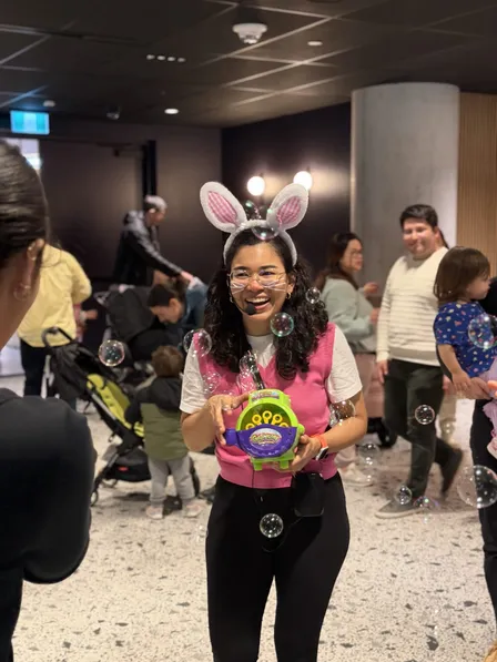 Karla, wearing bunny ears, laughs while holding a bubble machine as families play among floating bubbles.