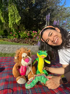 Ms. Karla smiles outdoors on a red tartan blanket, posing with a lion and dragon stuffed animal on a sunny day with flowers and trees in the background.