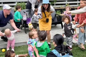 Karla in a yellow pinafore and blue bucket hat smiles while interacting with children outdoors, surrounded by parents and caregivers.
